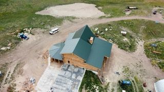 Aerial view of a house with a green roof on a dirt lot, surrounded by construction materials.