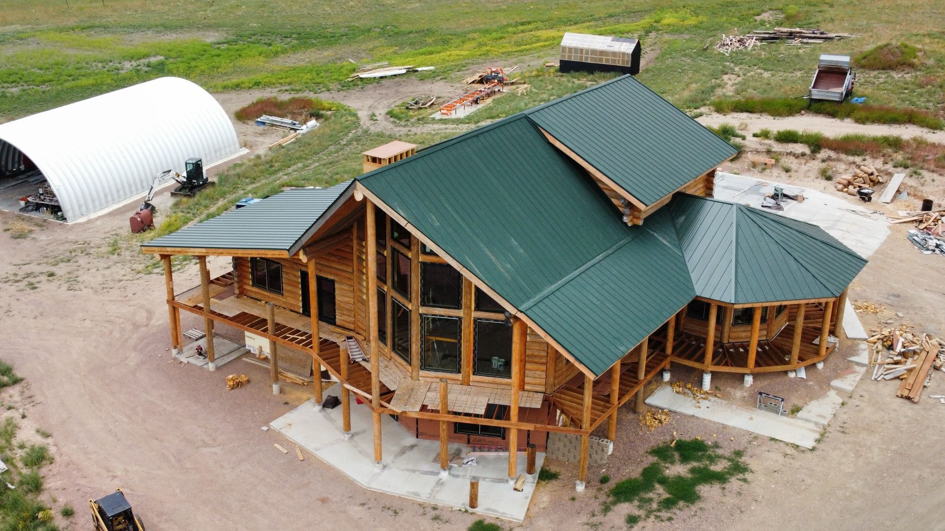 Aerial view of a large wooden house with green metal roofs in a rural landscape.