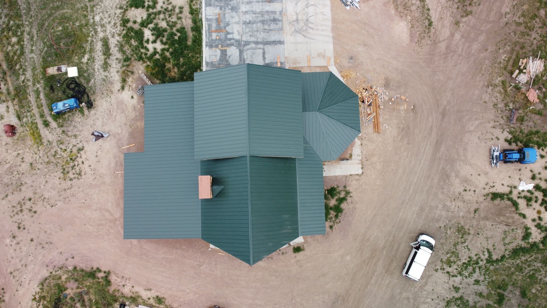 Aerial view of a green-roofed house with dirt driveway and parked vehicles nearby