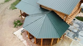 Aerial view of a log cabin with green metal roofs and a round porch on a dirt lot.