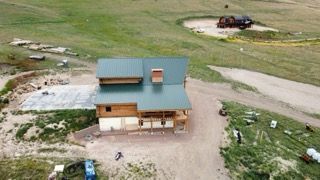 Aerial view of a small house with a green roof in a rural dirt lot, with a truck and open land nearby