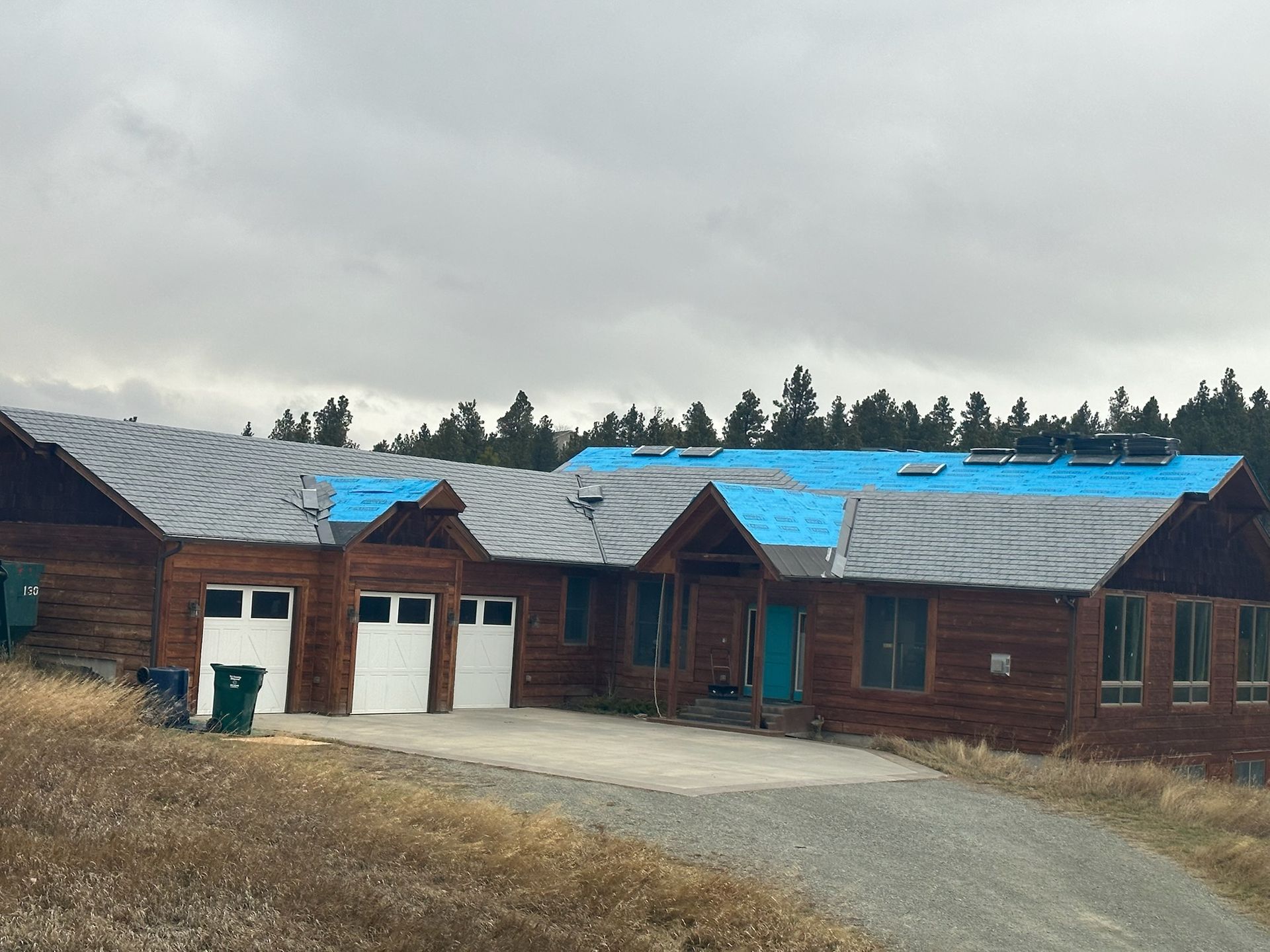 Rustic wooden buildings with blue and gray roofs along a gravel driveway under a cloudy sky