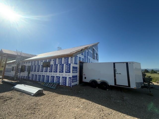 Partially built house wrapped in blue-and-white house wrap, with a white trailer parked beside it under a bright sky