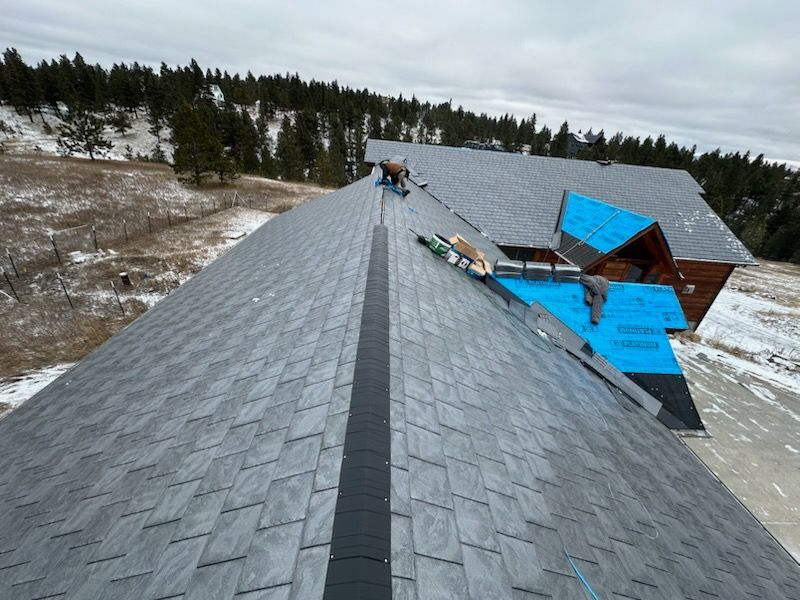 Roof under construction with gray shingles, blue tarp, and workers on a snowy hillside.