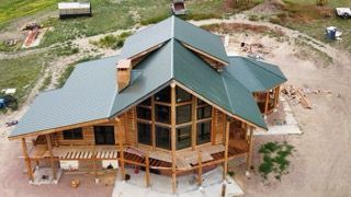 Aerial view of a large wooden house with a green metal roof on a dirt lot under construction
