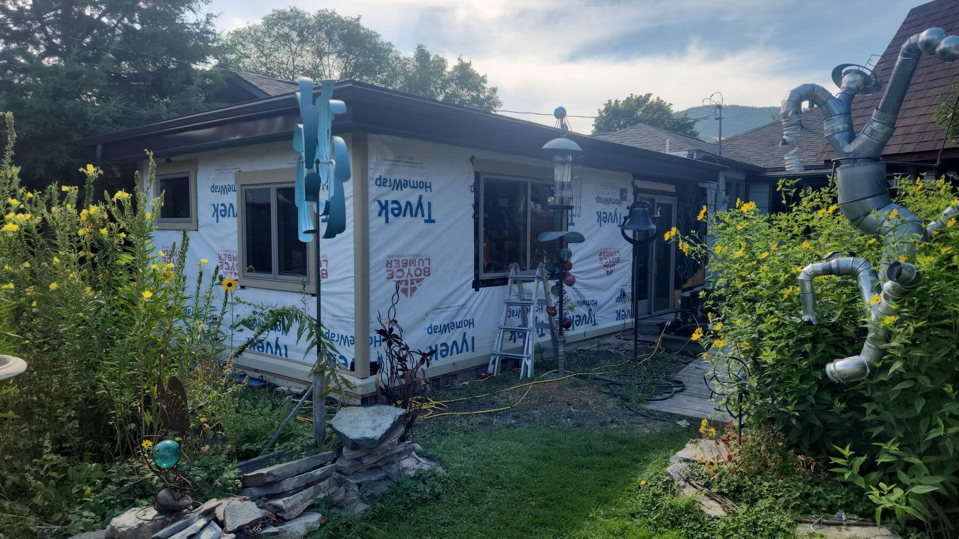 A house under construction with Tyvek wrap; surrounded by overgrown garden and art sculptures.