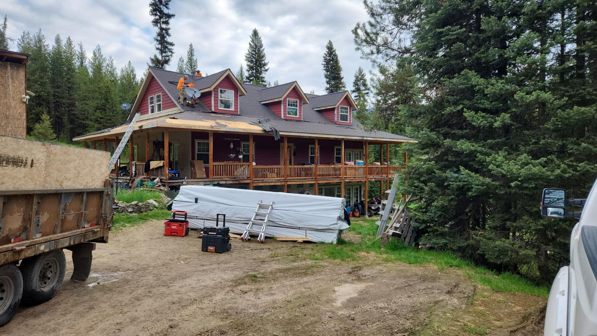 House under roof repair with workers, surrounded by trees, dirt ground. Truck and supplies in foreground.