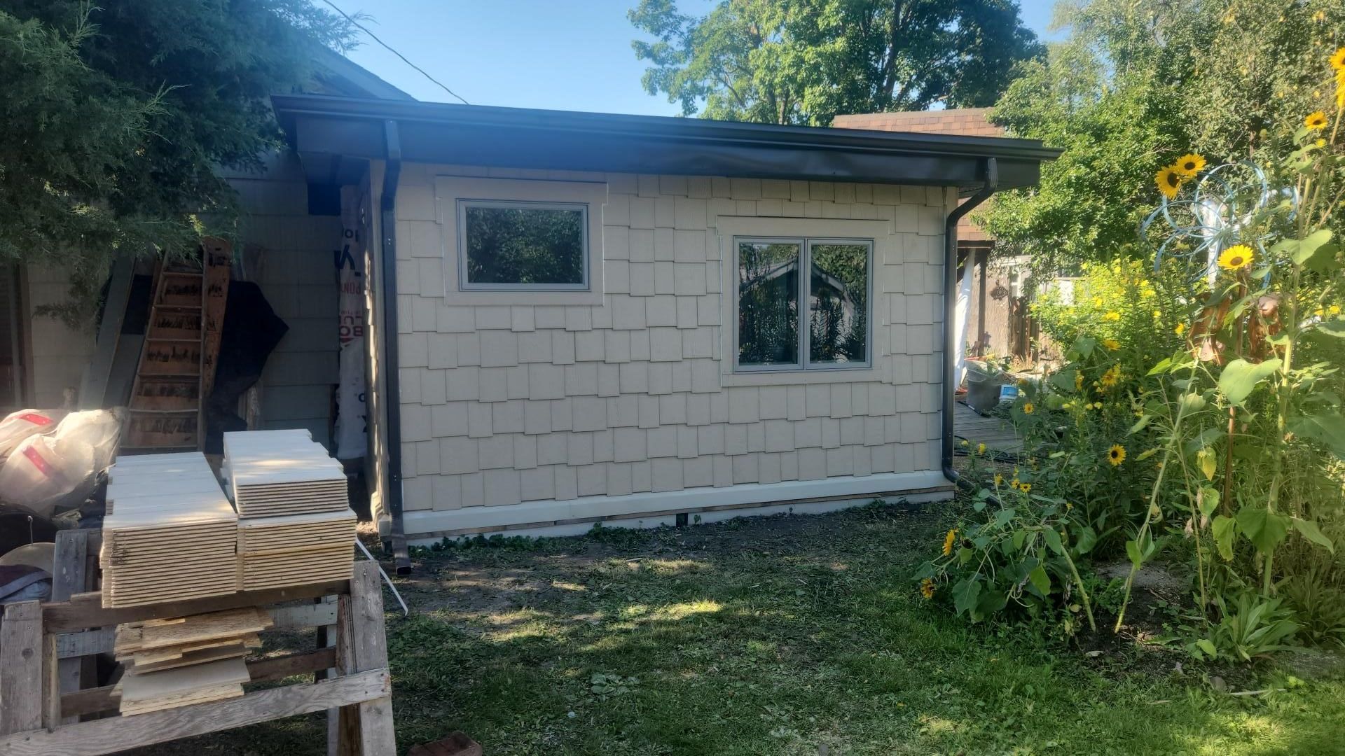 Beige shingled shed with two windows, overgrown grass, sunflowers. Sunny day.