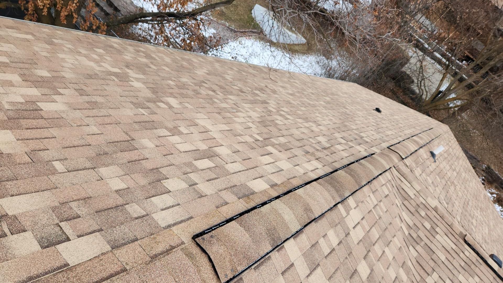 Brown asphalt shingle roof on a house, with a snowy yard and trees visible in the background.