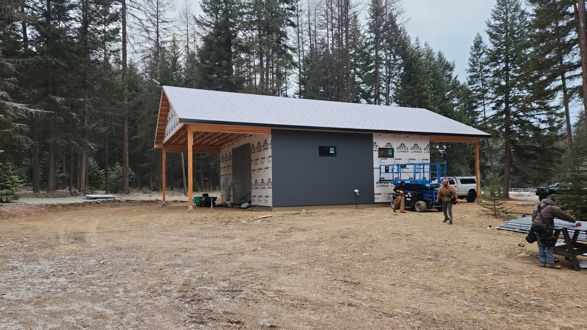 Construction site, gray building with wood overhang, surrounded by trees. Two people, a dog and a truck present.