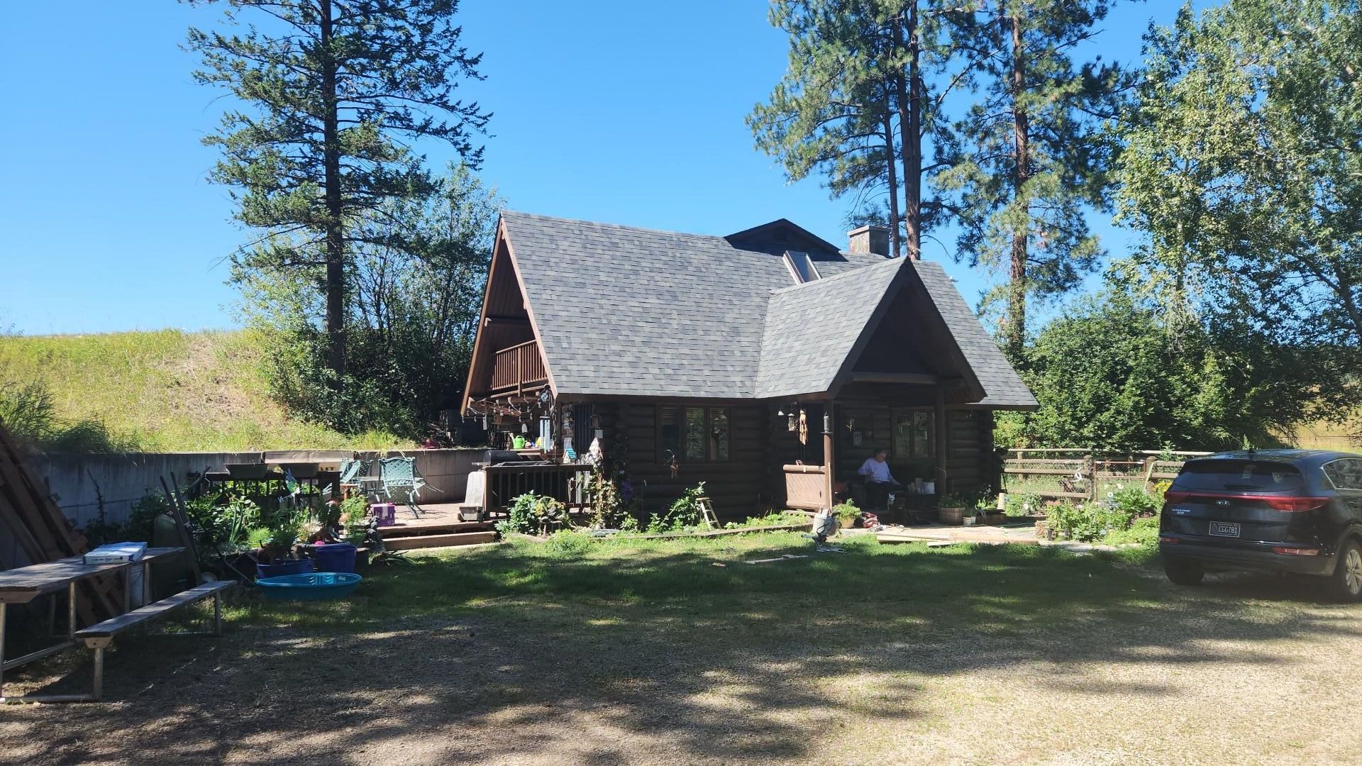 Log cabin with dark roof and porch, surrounded by greenery and a vehicle on a sunny day.