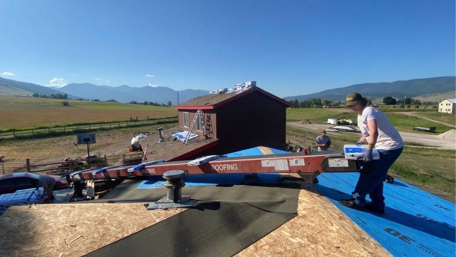 People installing roofing on a building in a rural setting, sunny day, mountains in the background.