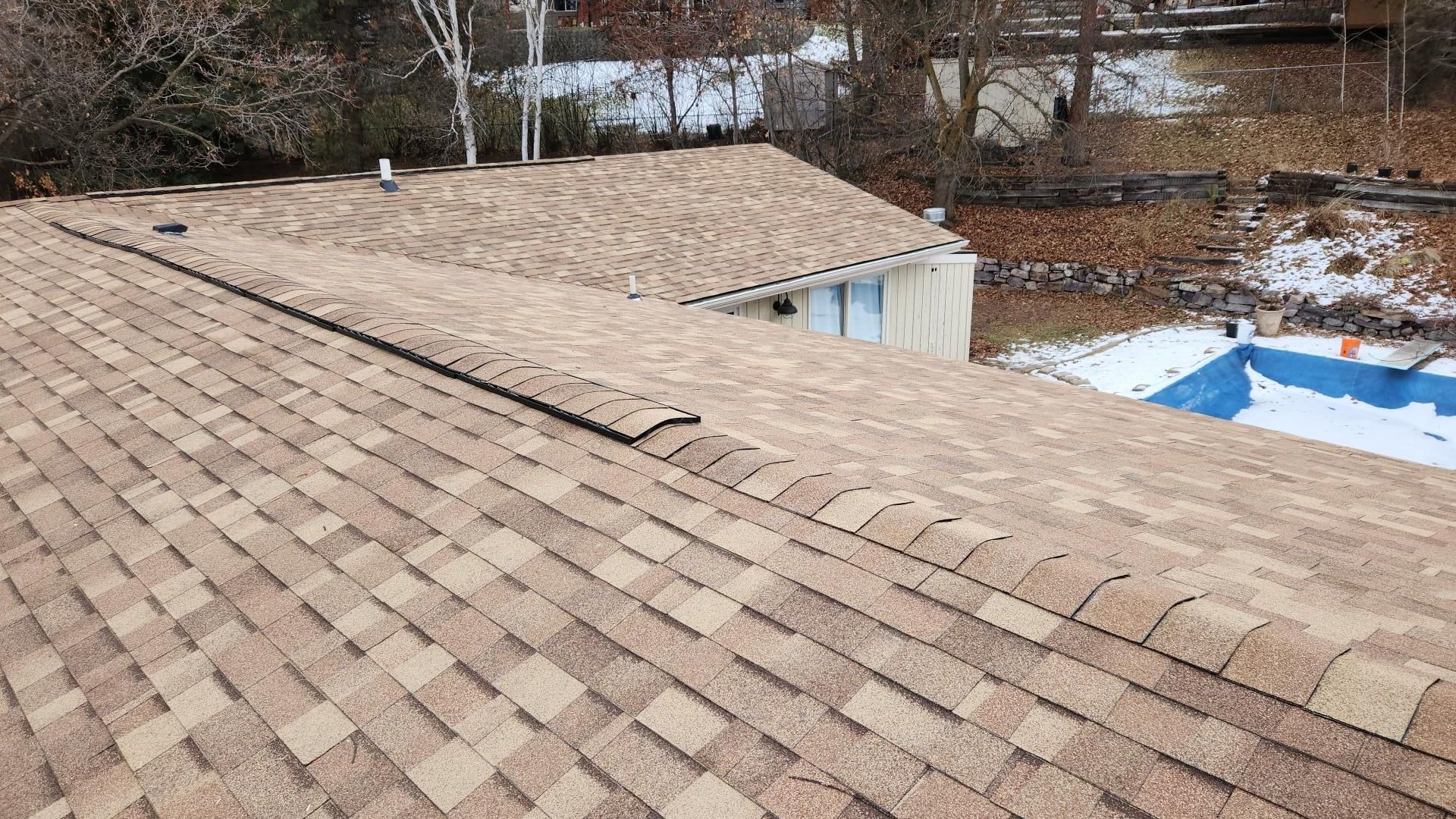 Brown shingled roof of a house with a pool in the background and a partly snowy landscape.