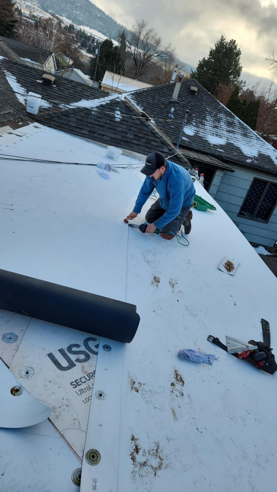 Person on a rooftop cuts material while installing roofing, partially covered in snow; mountains in the background.