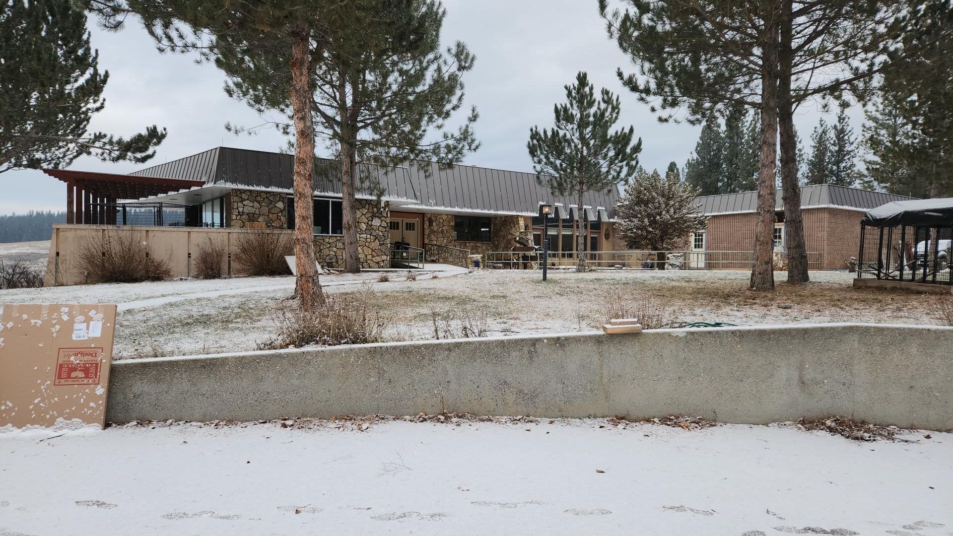 Snow-covered, one-story house with stone facade, under a cloudy sky. Trees and a concrete wall are in the foreground.