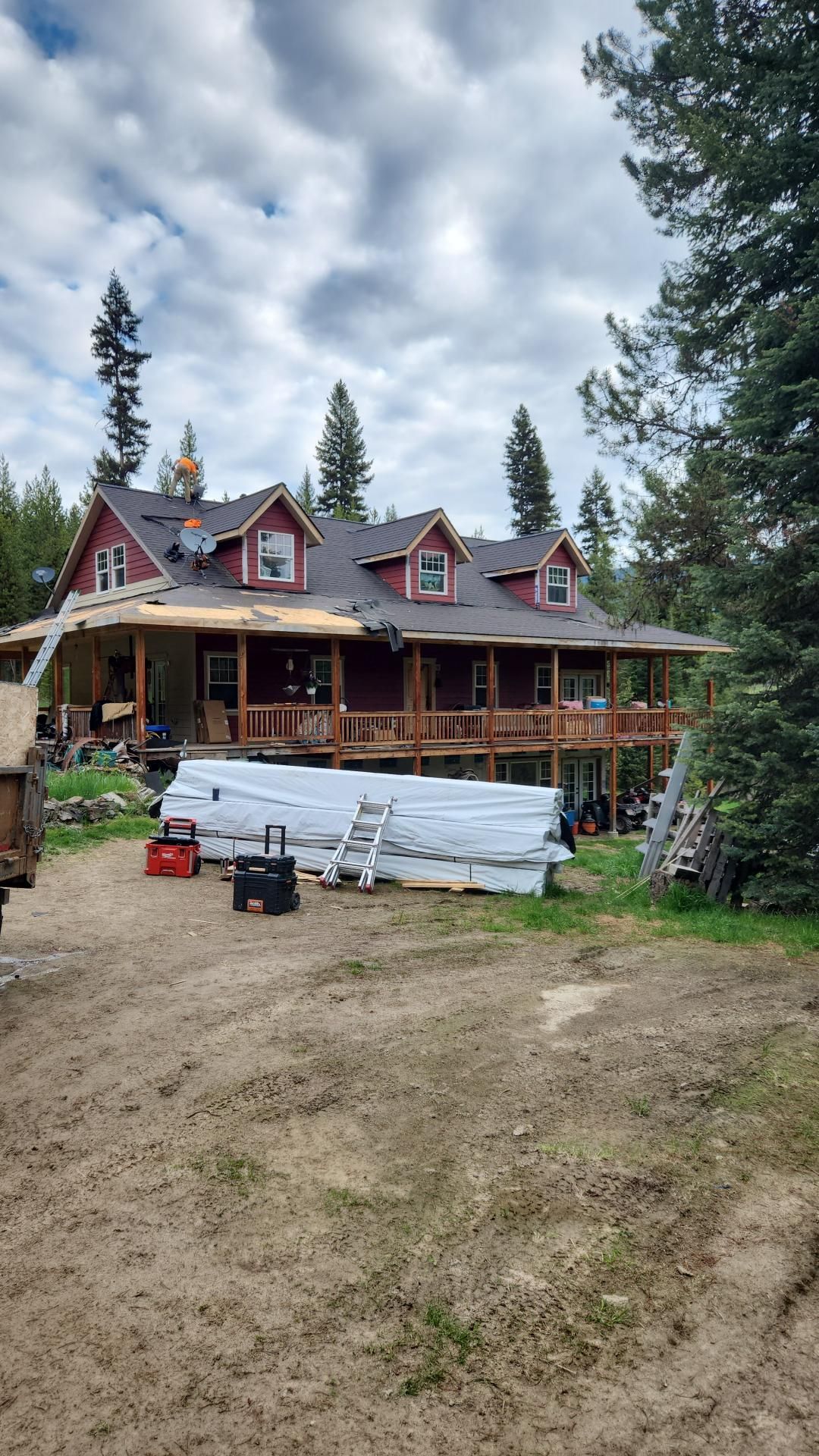 House under construction; red siding, wrap-around porch, brown roof, lumber and supplies on ground, forested setting.