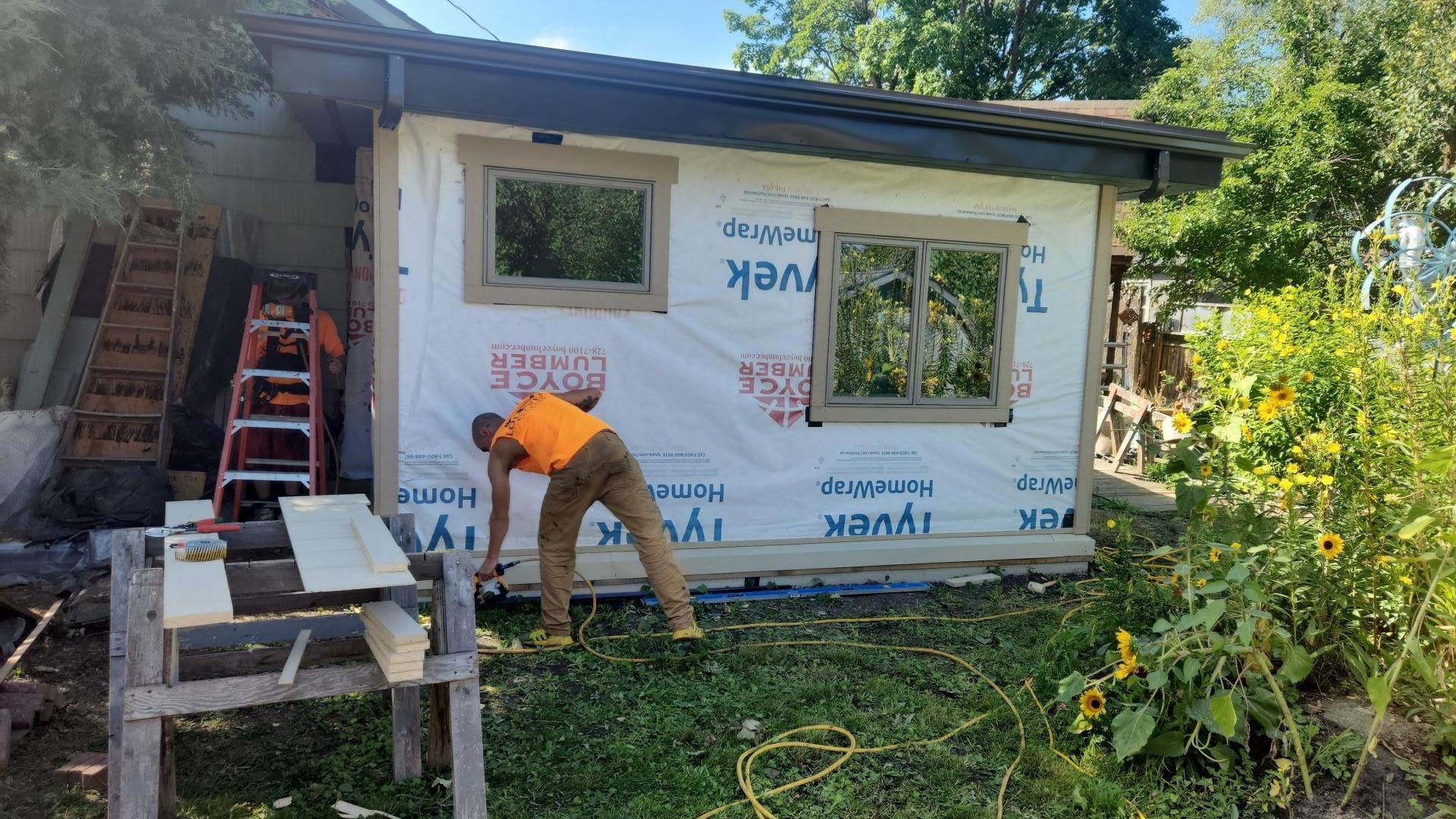 Man working on a small shed, attaching wood trim. Tyvek wrap, windows, and green yard.
