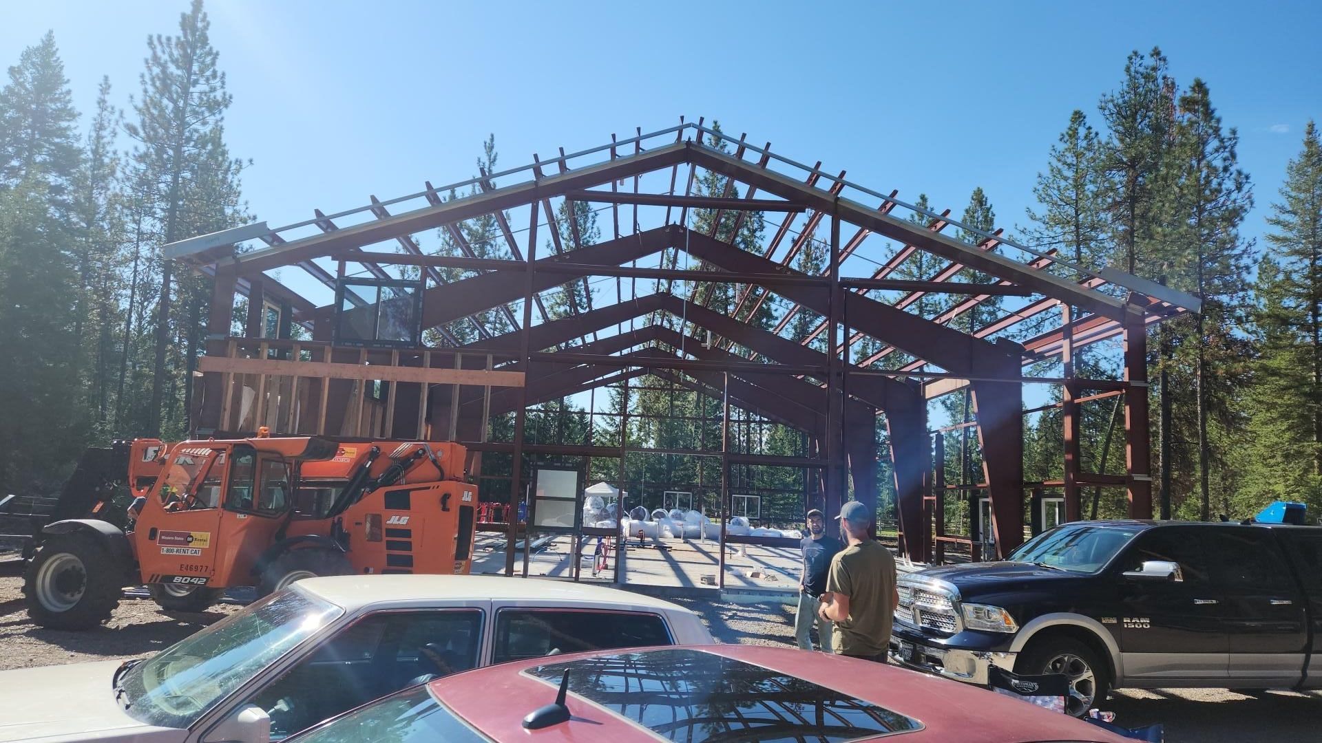 Construction of a metal framed building in a wooded area. People, orange lift, vehicles visible.