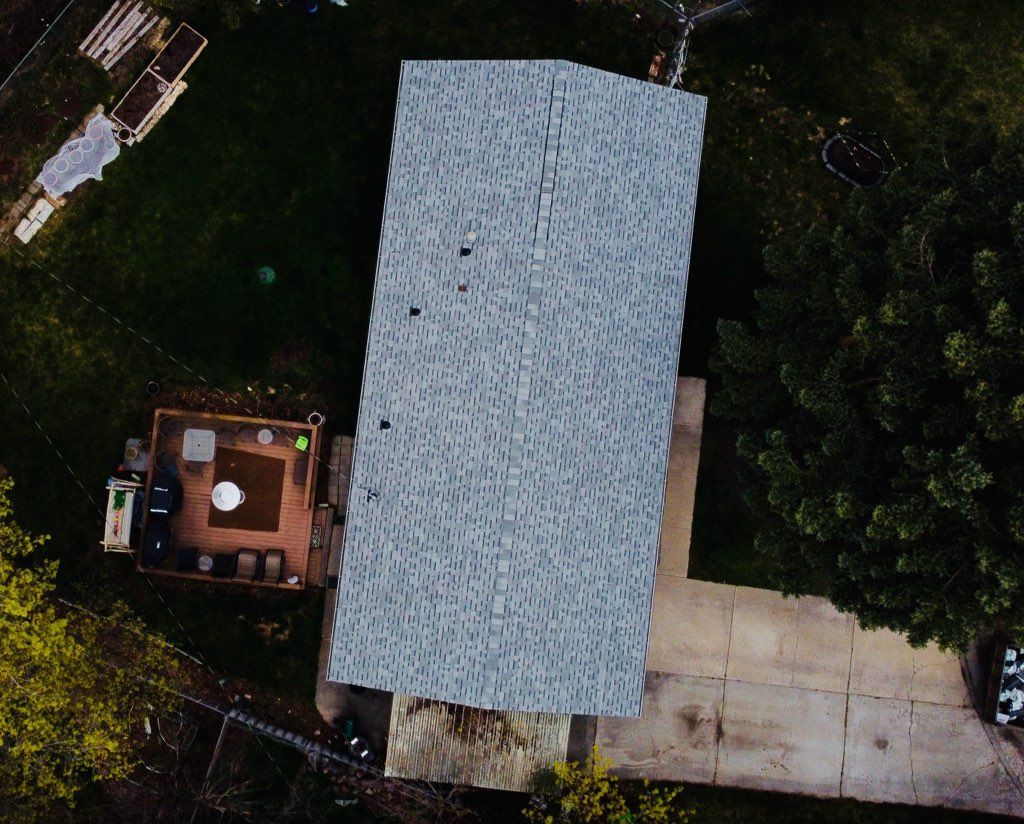 Aerial view of a house with a gray roof, adjacent wooden deck, and driveway surrounded by trees.