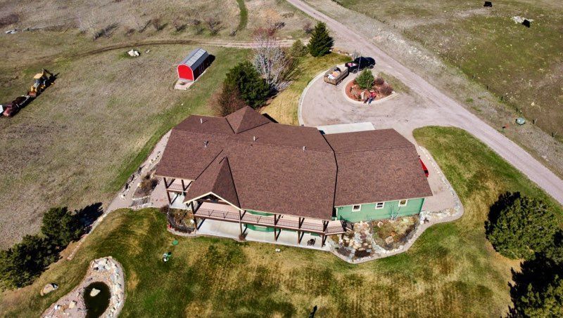 Aerial view of a brown-roofed house with a green side, surrounded by open grass and gravel driveways.
