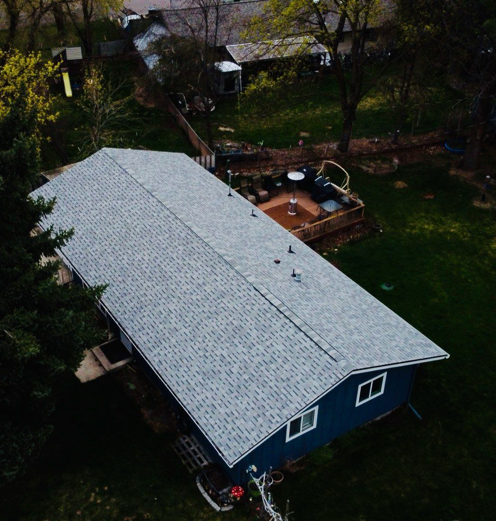 Aerial view of a blue house with a gray roof and a backyard deck with a hot tub surrounded by trees.