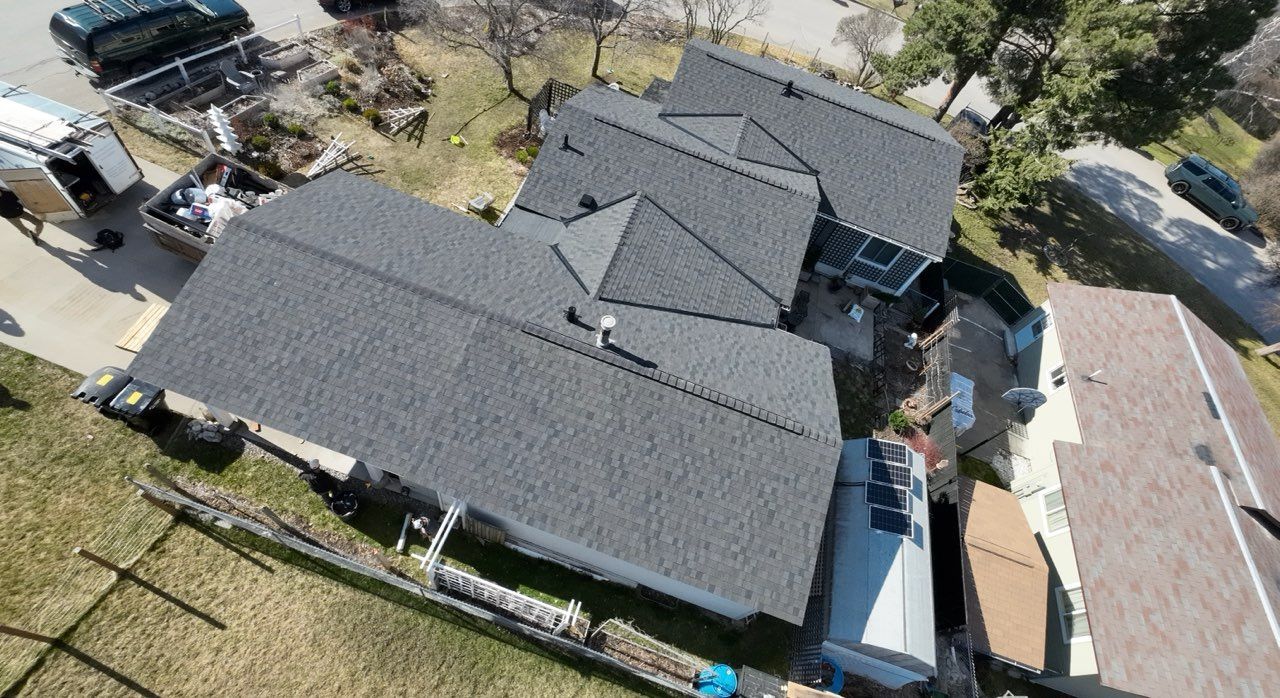 Aerial view of a gray-roofed house with a yard, driveway, and neighboring homes.
