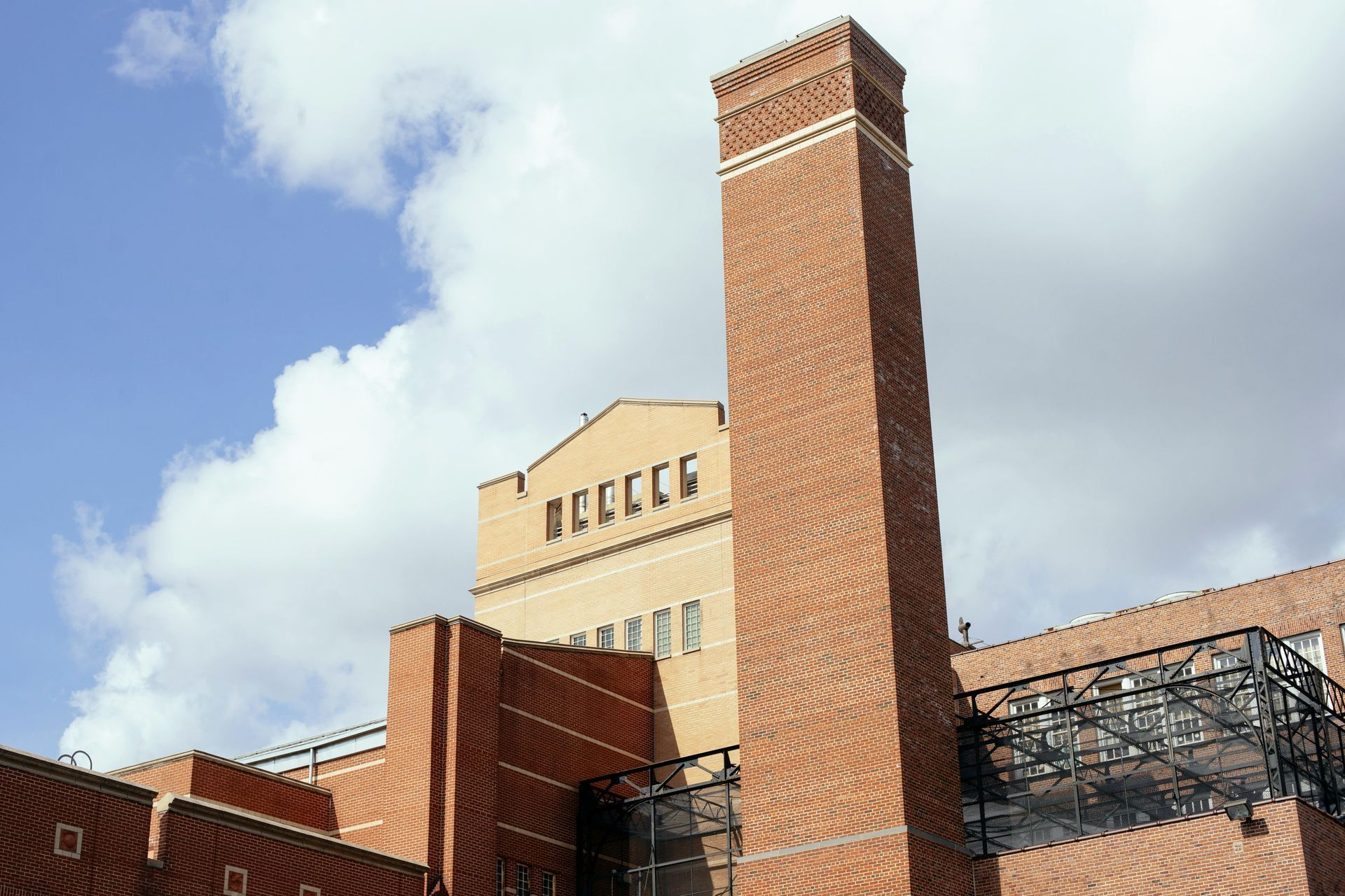 Tall red brick chimney against a blue sky with clouds, attached to a building with a light-colored facade.