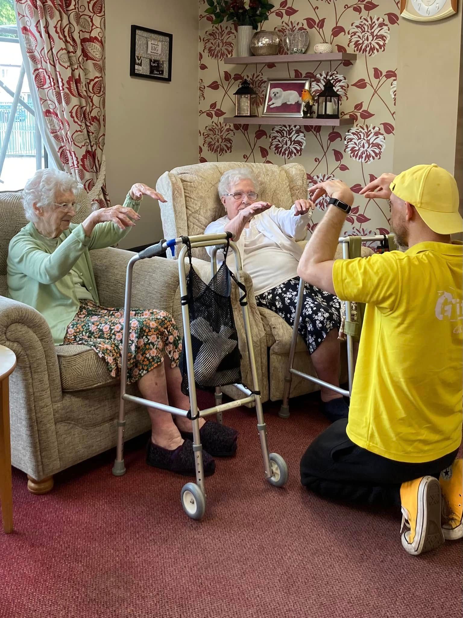 Care worker kneels to assist an elderly person in a living room while another person watches from a chair