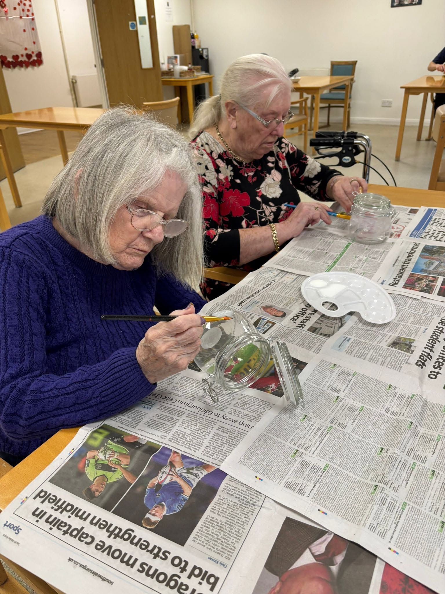 Two people painting at a table covered in newspaper in a bright community room.