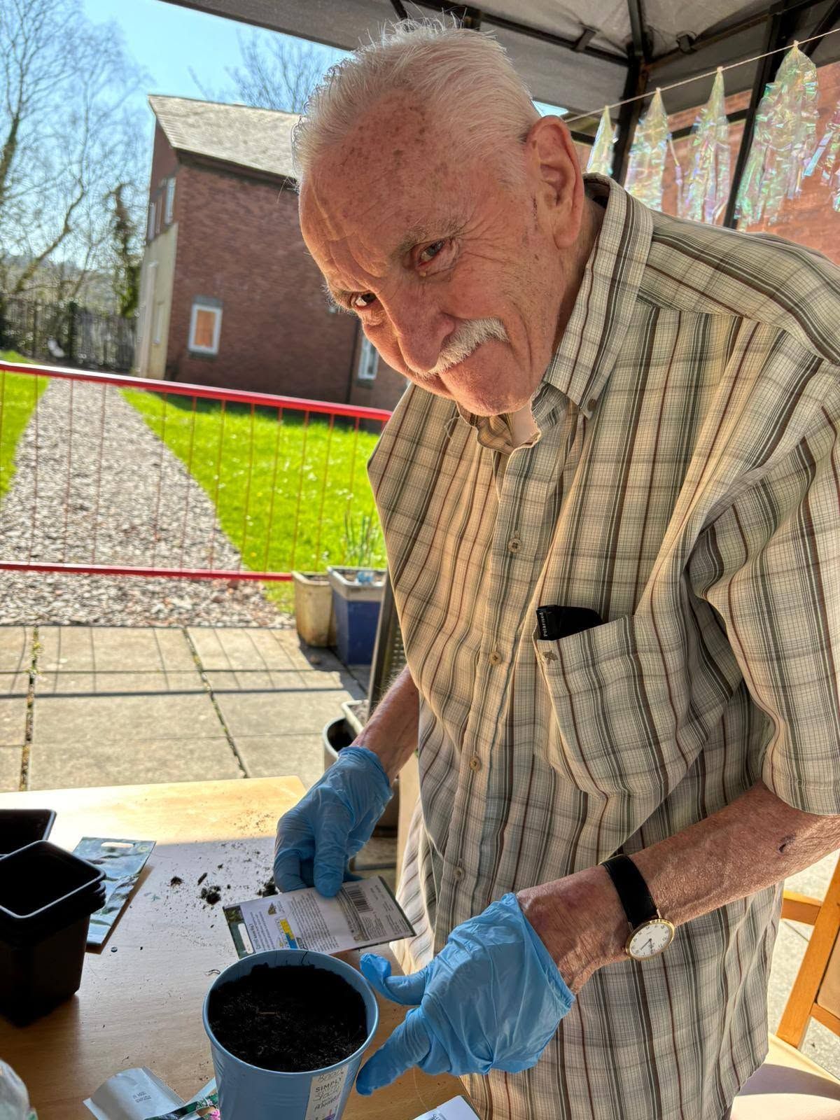 Elderly man in blue gloves pouring soil into a pot at an outdoor patio table.