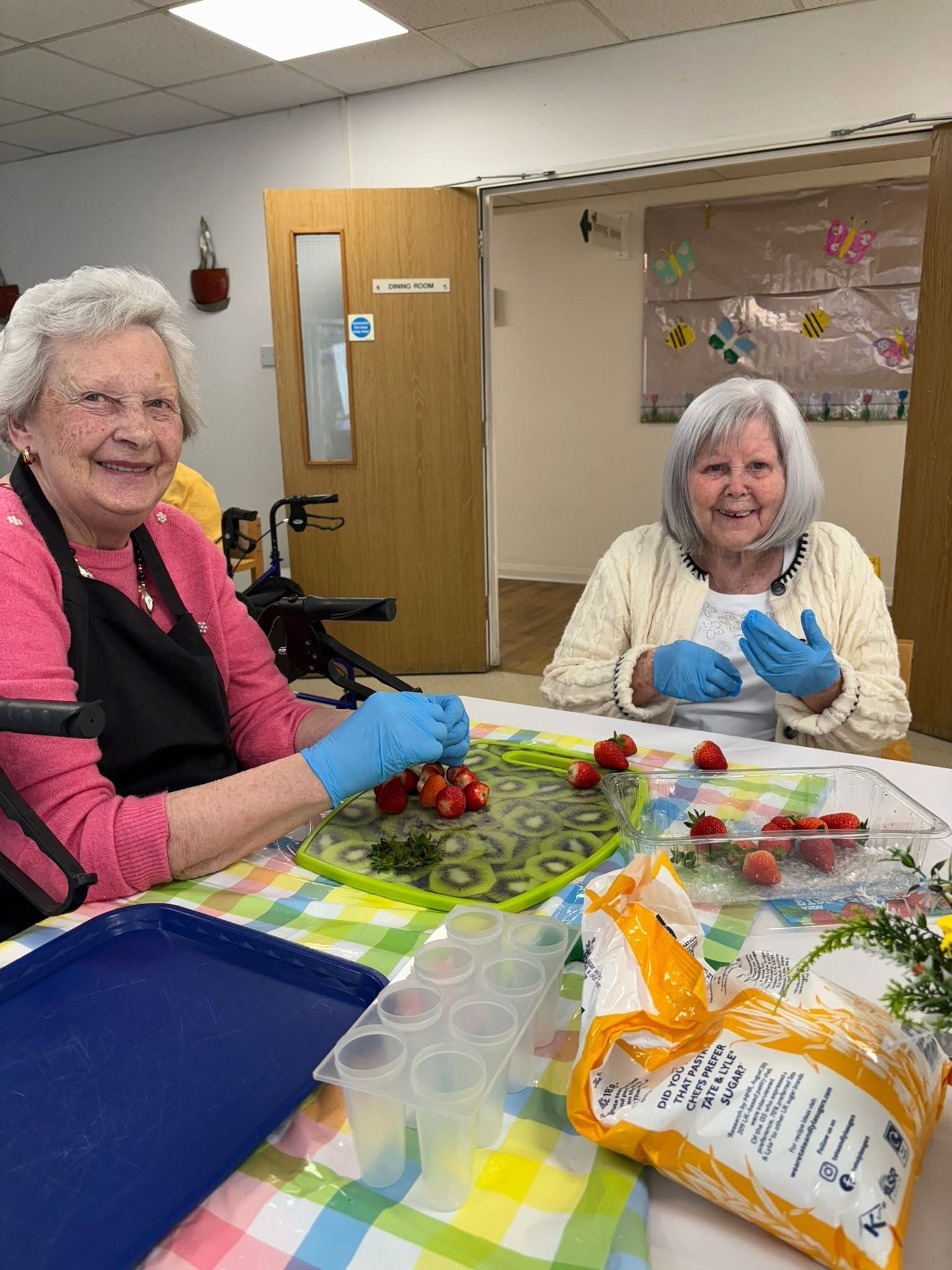 Two people in blue gloves prepare food at a table with lettuce, strawberries, and an open snack bag.