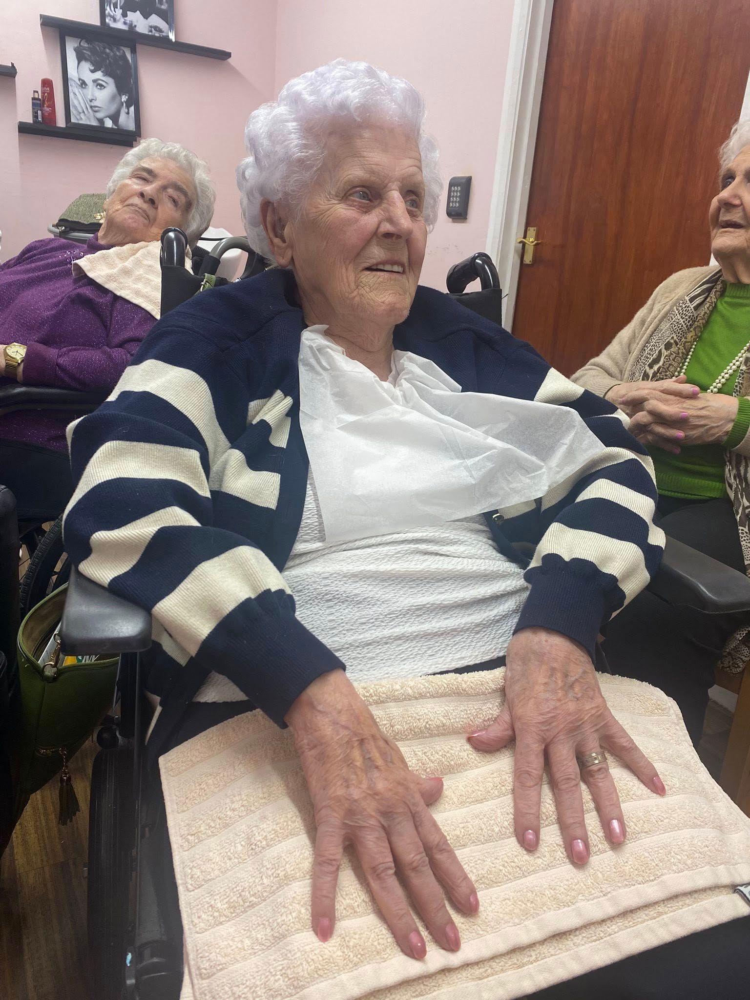 Three elderly women seated indoors, with the center woman resting her hands on a blanket.