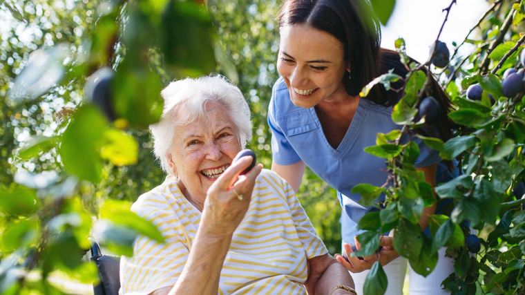 Two women smiling among grapevines, one offering grapes to the other outdoors.
