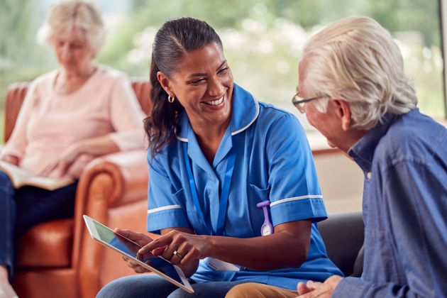 Nurse in blue scrubs smiling and talking with an older adult in a waiting room, clipboard in hand