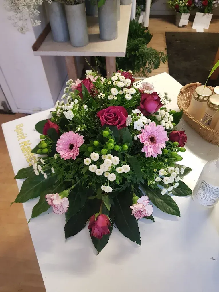 Hjerteformet blomsterarrangement med gule solsikker, røde gerberaer og orange roser, placeret på en træbænk.