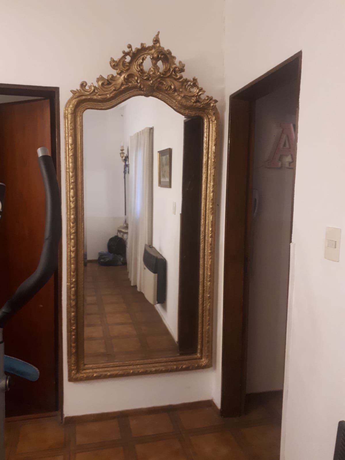 Espejo ornamentado con marco dorado en un pasillo, que refleja un largo corredor y una puerta.