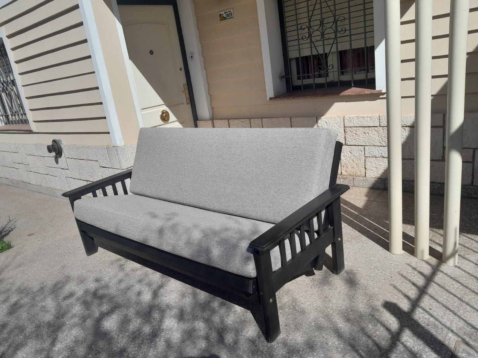 Banco acolchado gris en un patio soleado junto a una casa beige con sombras a rayas.