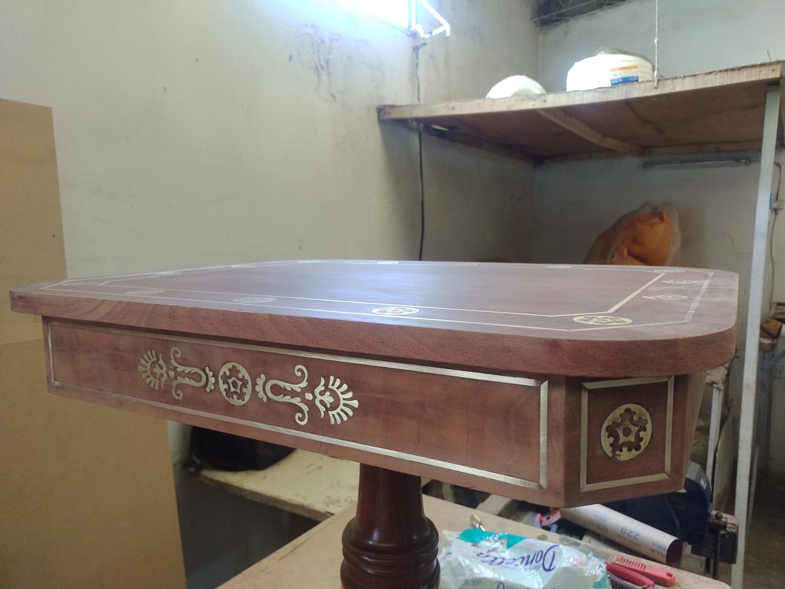 Mesa de comedor de madera con tallas florales en una habitación pequeña, cerca de una litera y una lámpara de pared.