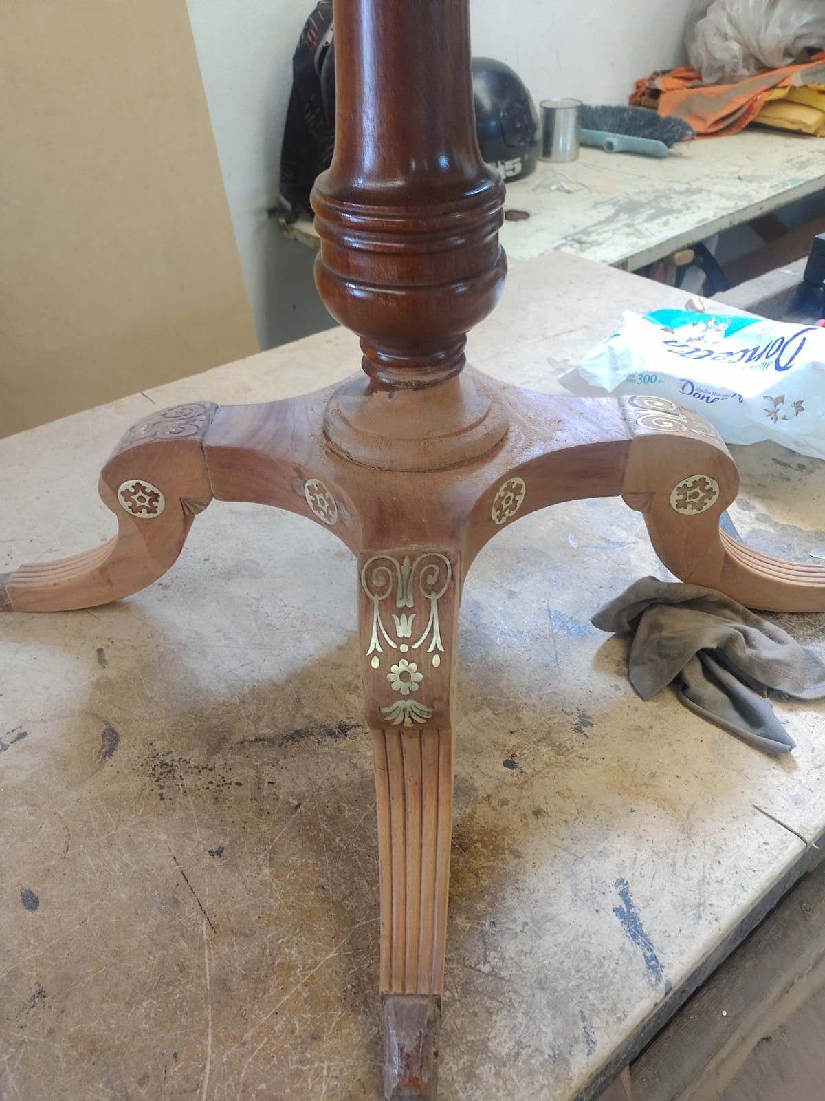 Base de mesa de madera con patas de trípode talladas sobre un banco de trabajo, con herramientas al fondo.