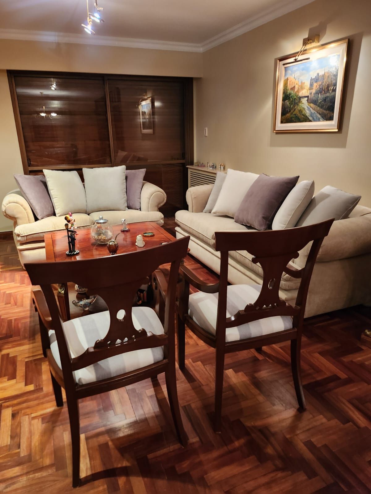 Acogedora sala de estar con sillas de madera, sofá beige, mesa de centro y cuadro de paisaje enmarcado.