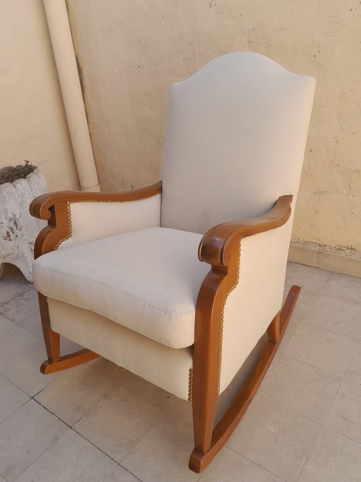 Sillón tapizado en blanco con brazos de madera y respaldo curvo, colocado en un patio embaldosado contra una pared beige.