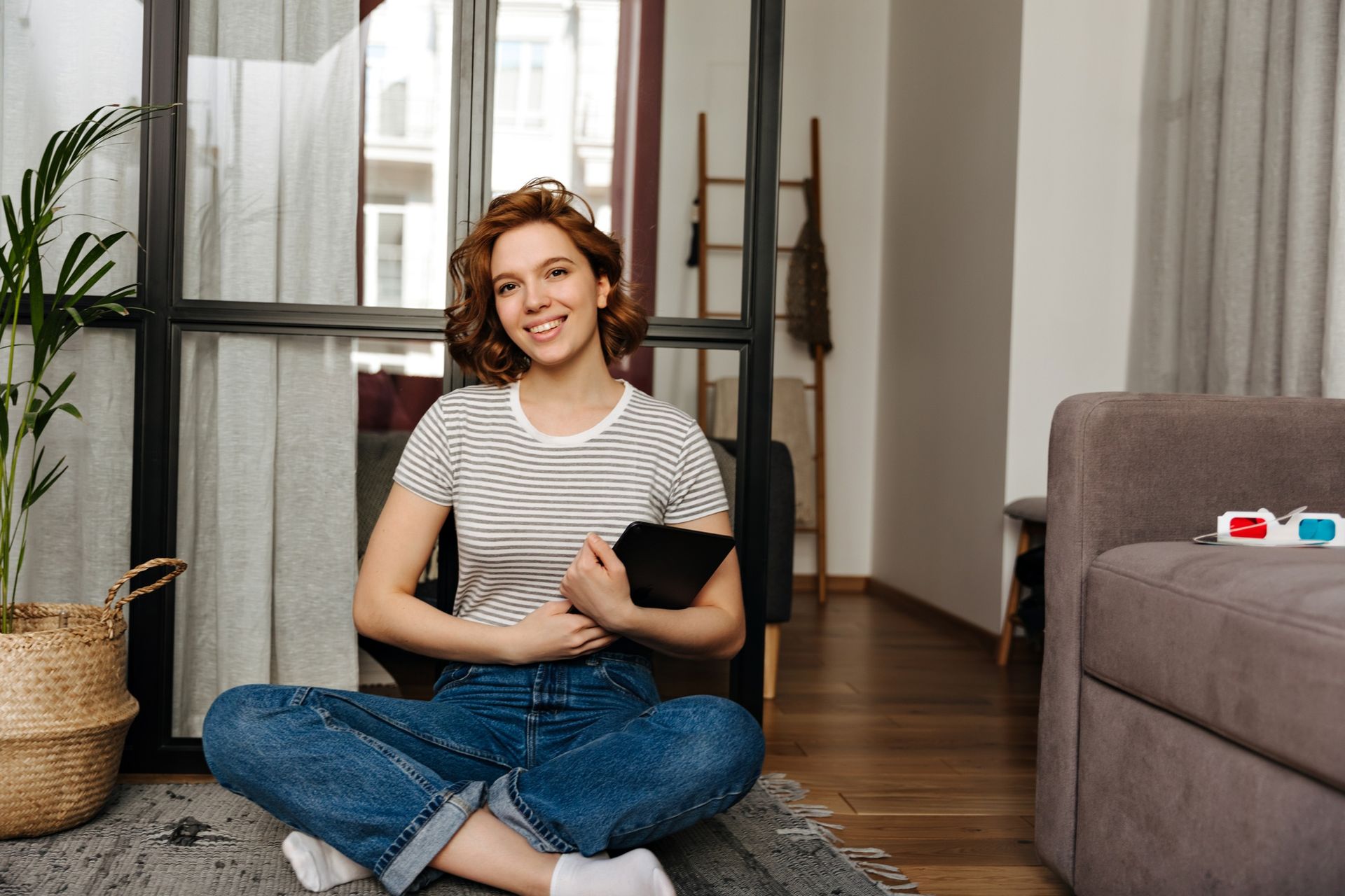 Smiling person sitting cross-legged on a rug holding a black cat in a bright living room