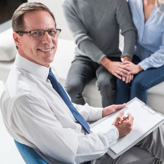 Doctor in white shirt and blue tie taking notes during a counseling session with seated clients