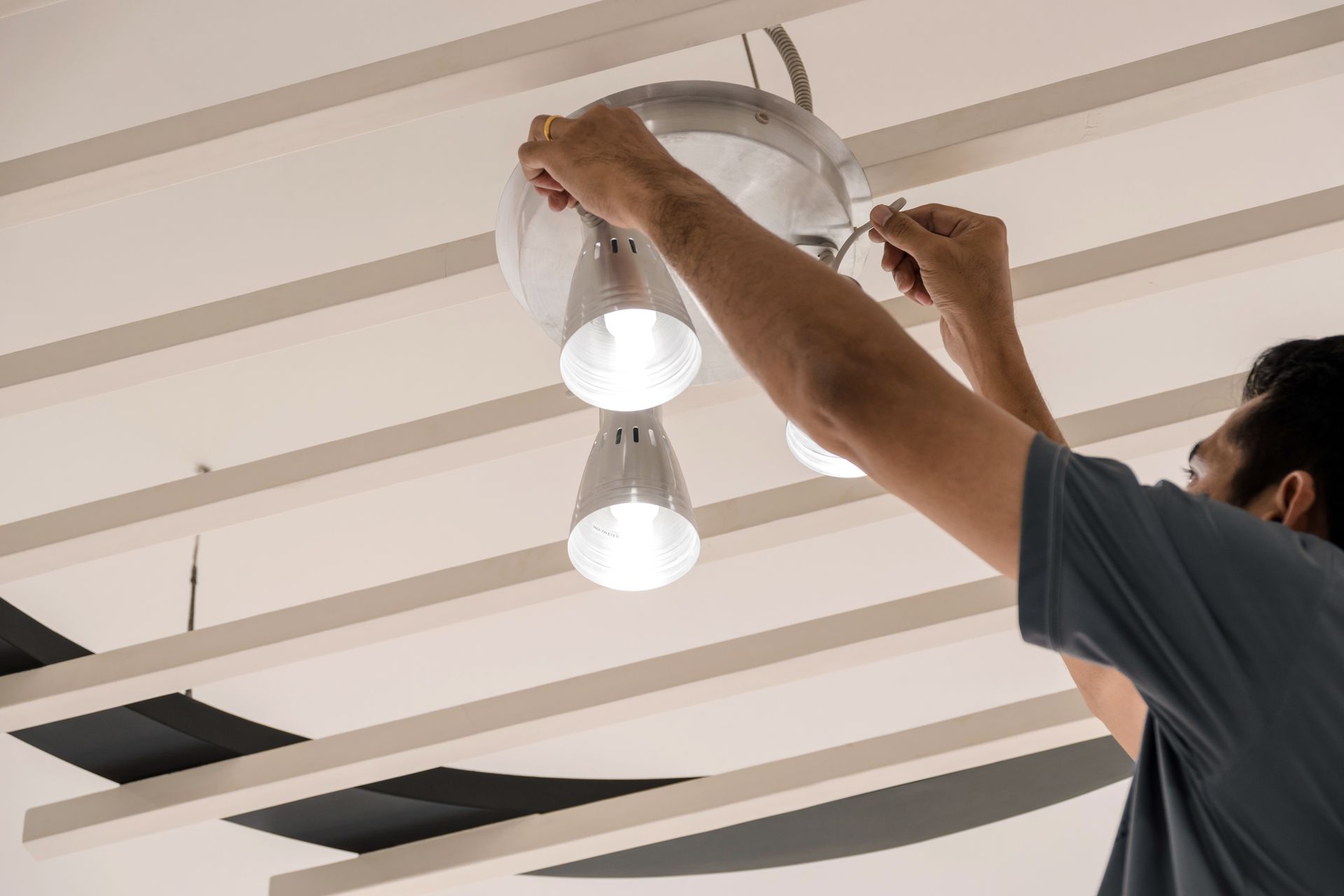 Persona instalando una lámpara de techo con tres focos; techo blanco con vigas.