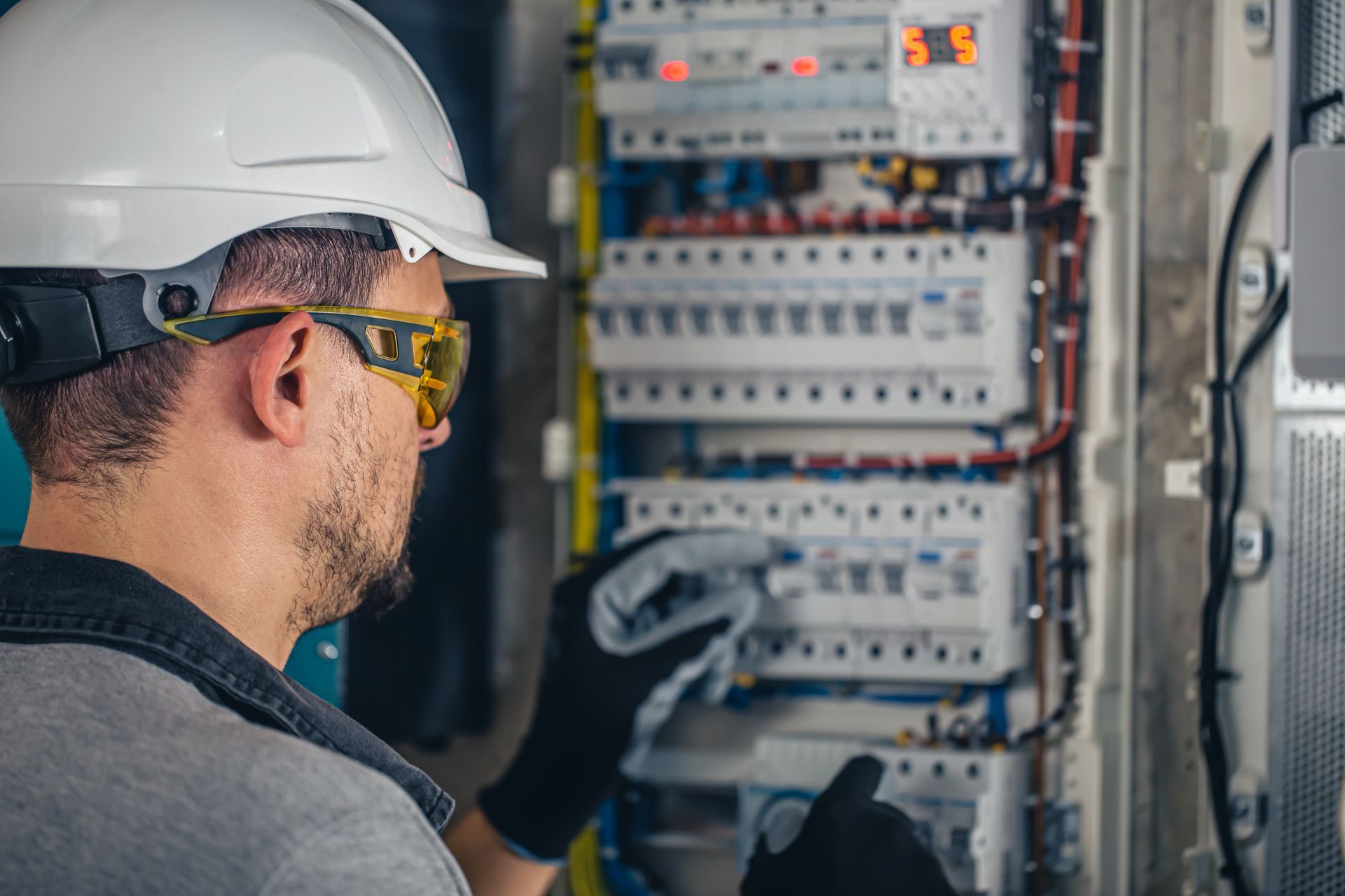 Electricista con casco y gafas de seguridad trabajando en un panel de interruptores automáticos.