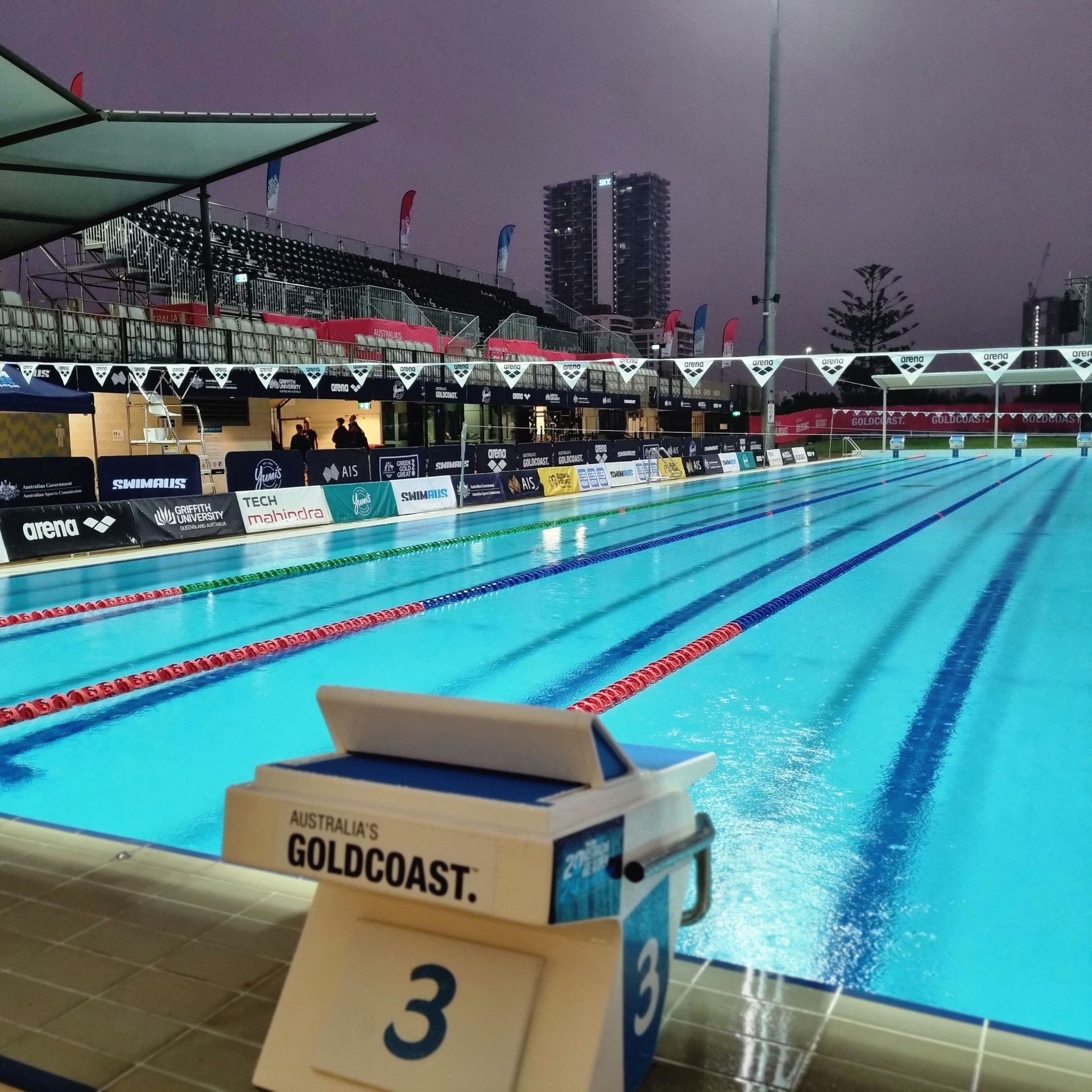 Swimming pool at Australia's Gold Coast, with a starting block in foreground, empty lanes, and stadium seating.