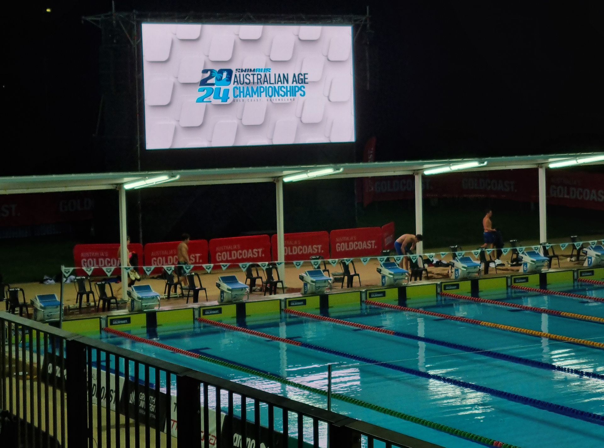 Swimming pool at night with starting blocks, scoreboard, and swimmers.