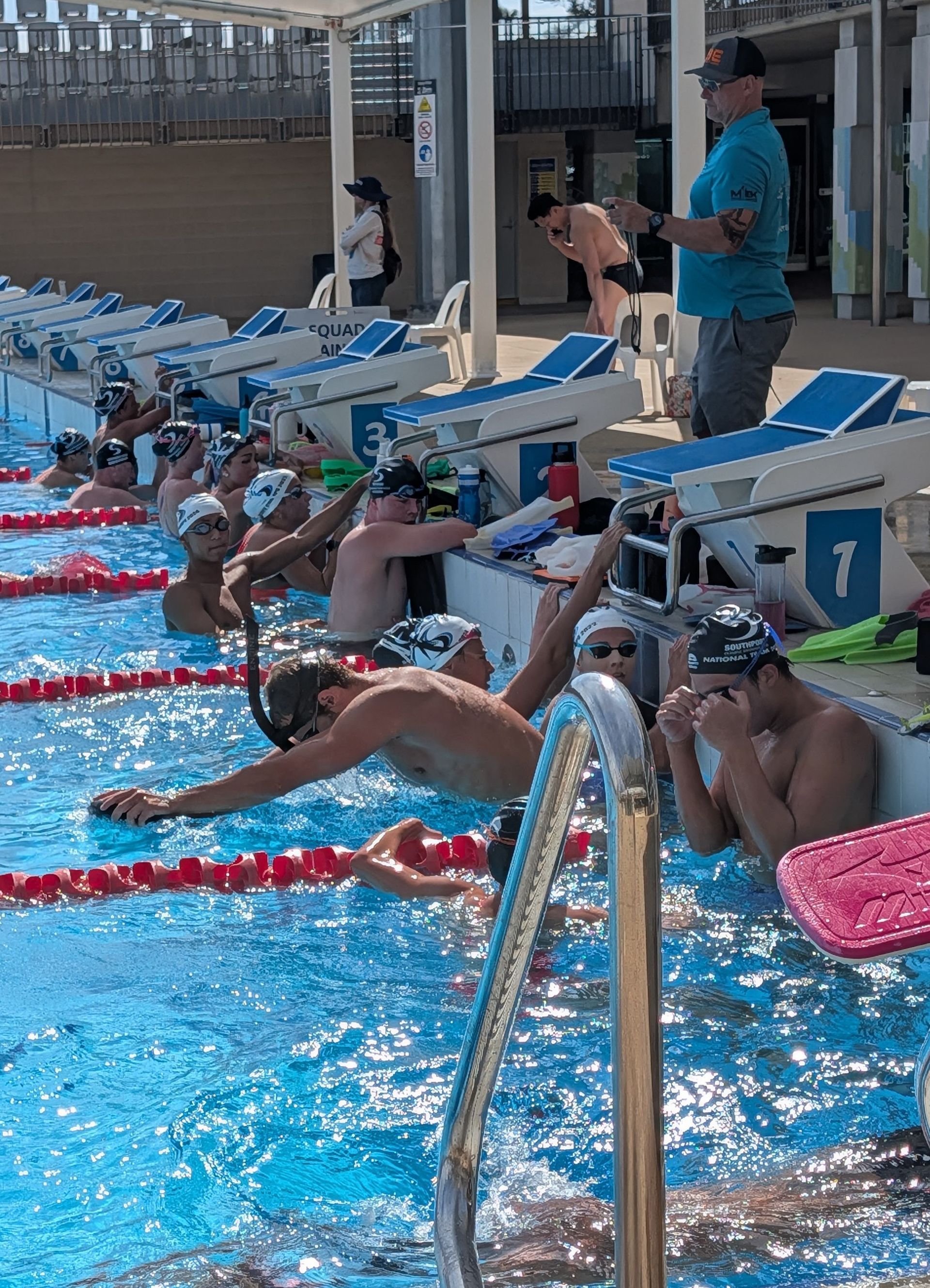 Swimmers in red and black suits poised on starting blocks at a pool, ready to dive.