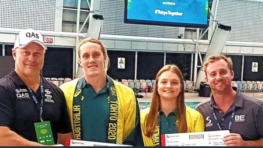 Group of four people; two athletes with medals, pose in front of Olympic signage at a pool.