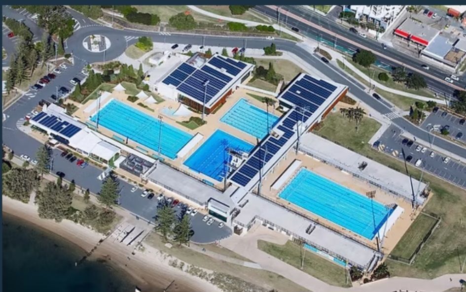 Aerial view of a public swimming pool complex with multiple blue pools, surrounded by walkways and solar panels.